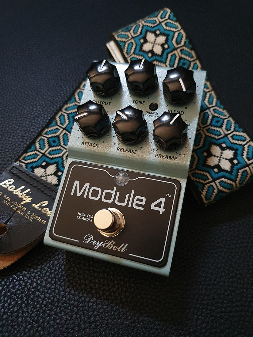 DryBell Module 4 guitar compressor alongside a guitar strap.